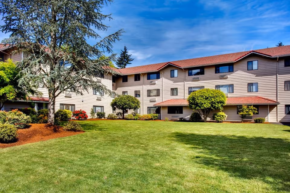 Exterior view of a multi-story retirement facility building with beige siding and red roof tiles, surrounded by a well-maintained lawn, trees, and shrubs under a blue sky with some clouds.