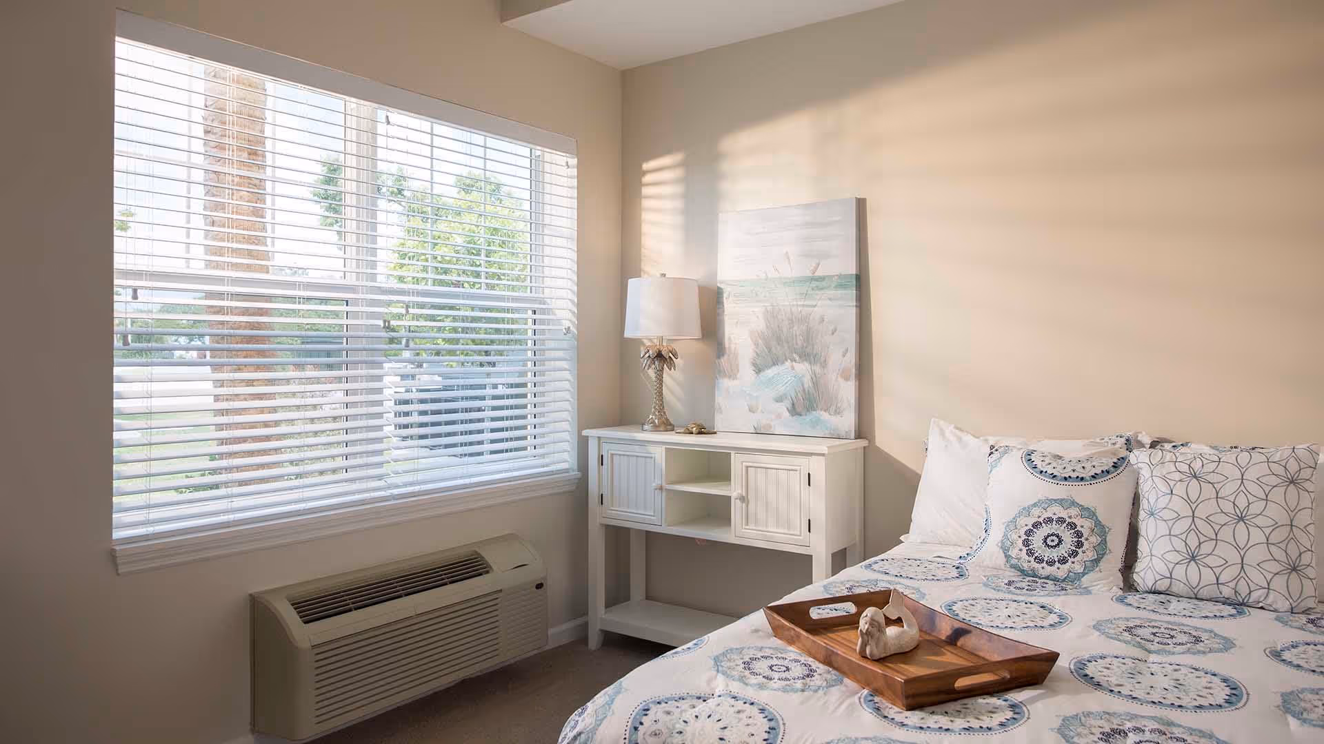 A bright bedroom with a large window covered by white blinds, a white air conditioning unit below the window, a white side table with a lamp and a beach-themed painting, and a bed with patterned blue and white bedding and pillows. A wooden tray with a small decorative item is placed on the bed.