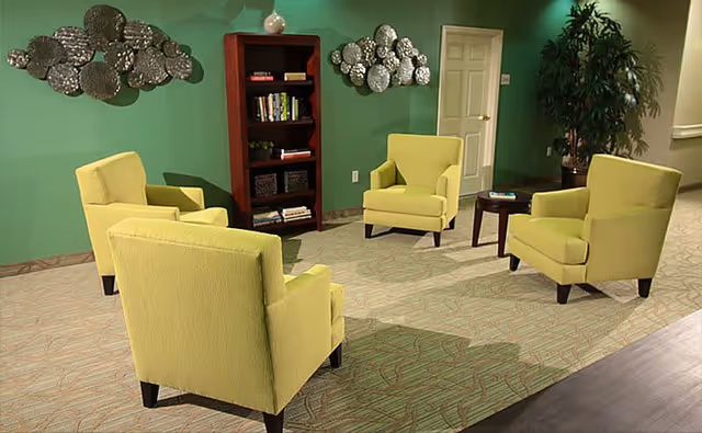 A cozy sitting area with four yellow armchairs arranged around a small dark wooden table. Behind the chairs is a green wall with two decorative metal wall art pieces and a wooden bookshelf filled with books and baskets. There is a door and a large potted plant in the corner.