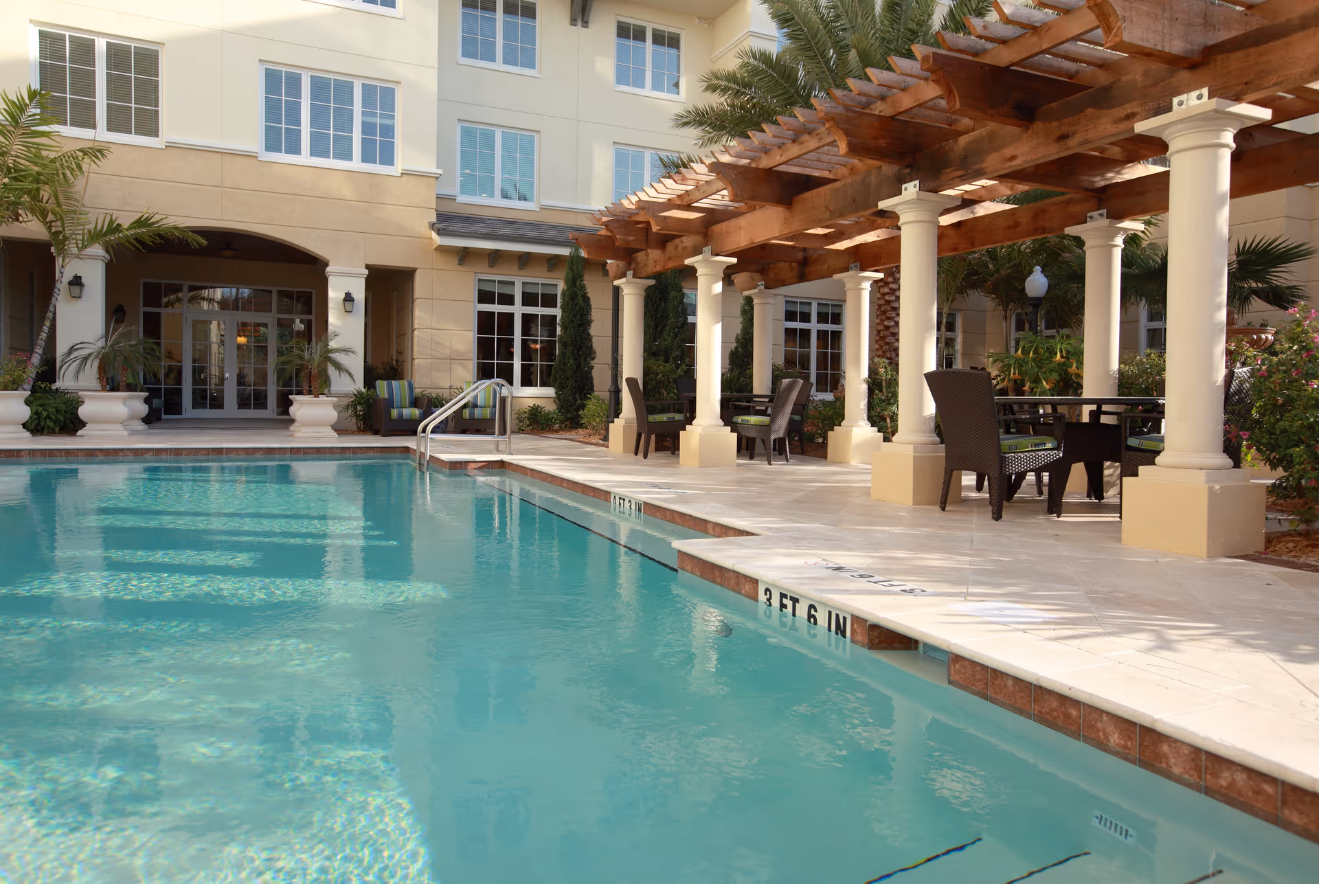 Outdoor swimming pool with a pergola, columns, and seating in front of a multi-story senior living building.