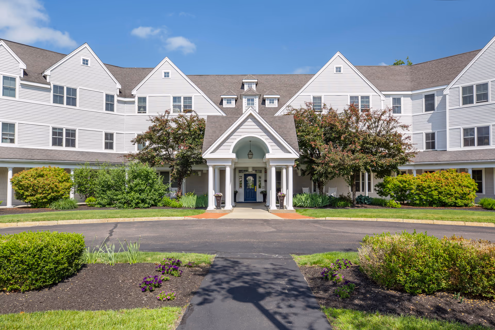 Front exterior view of a large, white, multi-story senior living facility building with a covered entrance supported by white columns. The building is surrounded by well-maintained landscaping including bushes, small trees, and flower beds under a clear blue sky.