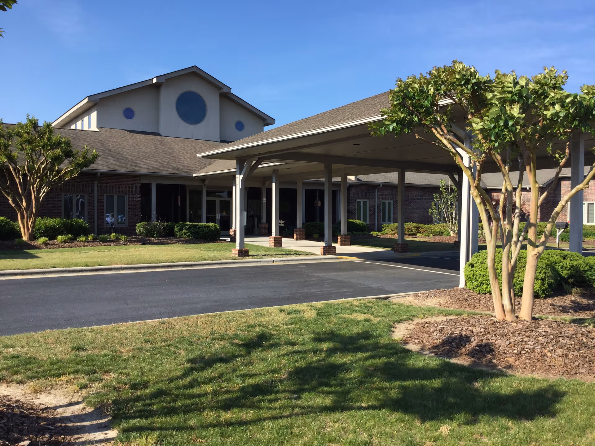 Front entrance of a brick senior living building with a covered porte-cochere and landscaped lawn.