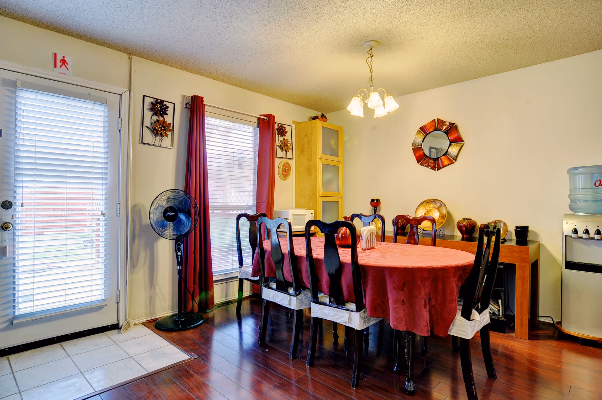 Dining room with a round table covered in a red tablecloth surrounded by chairs, a chandelier overhead, red curtains at the window, and a door to the left.