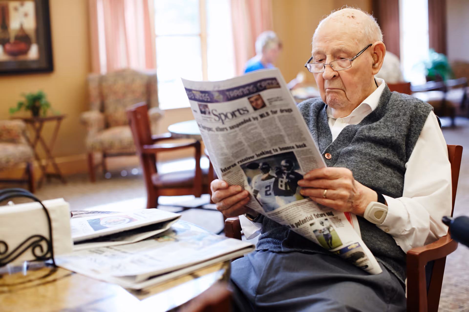 An elderly man wearing glasses and a gray vest is sitting in a chair reading a newspaper in a well-lit room with comfortable chairs and tables. Another person is visible in the background, also seated. The room has large windows with pink curtains and a warm, inviting atmosphere.