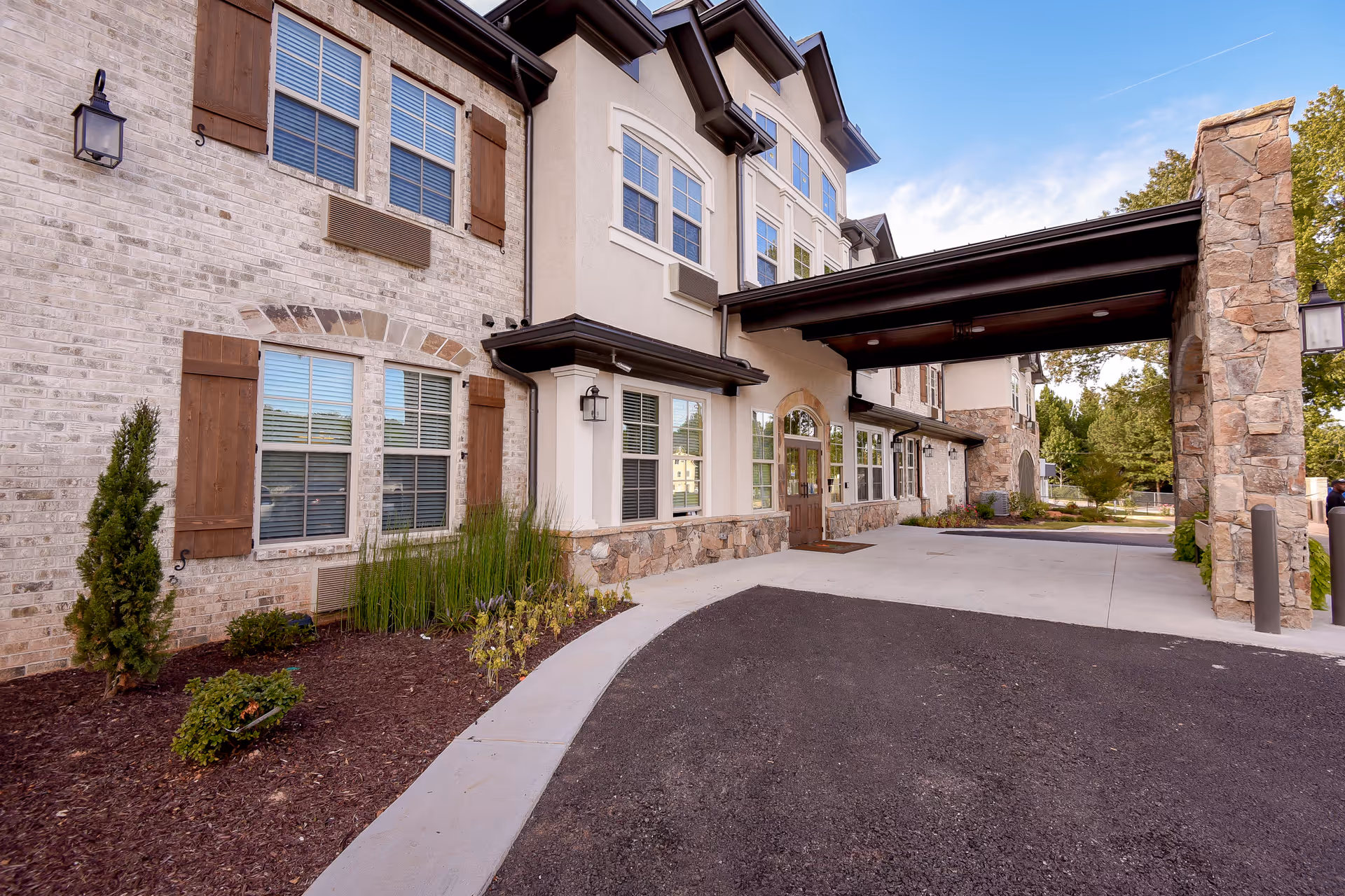 Covered entrance and driveway of a multi-story senior living building with stone and brick facade and landscaped beds.