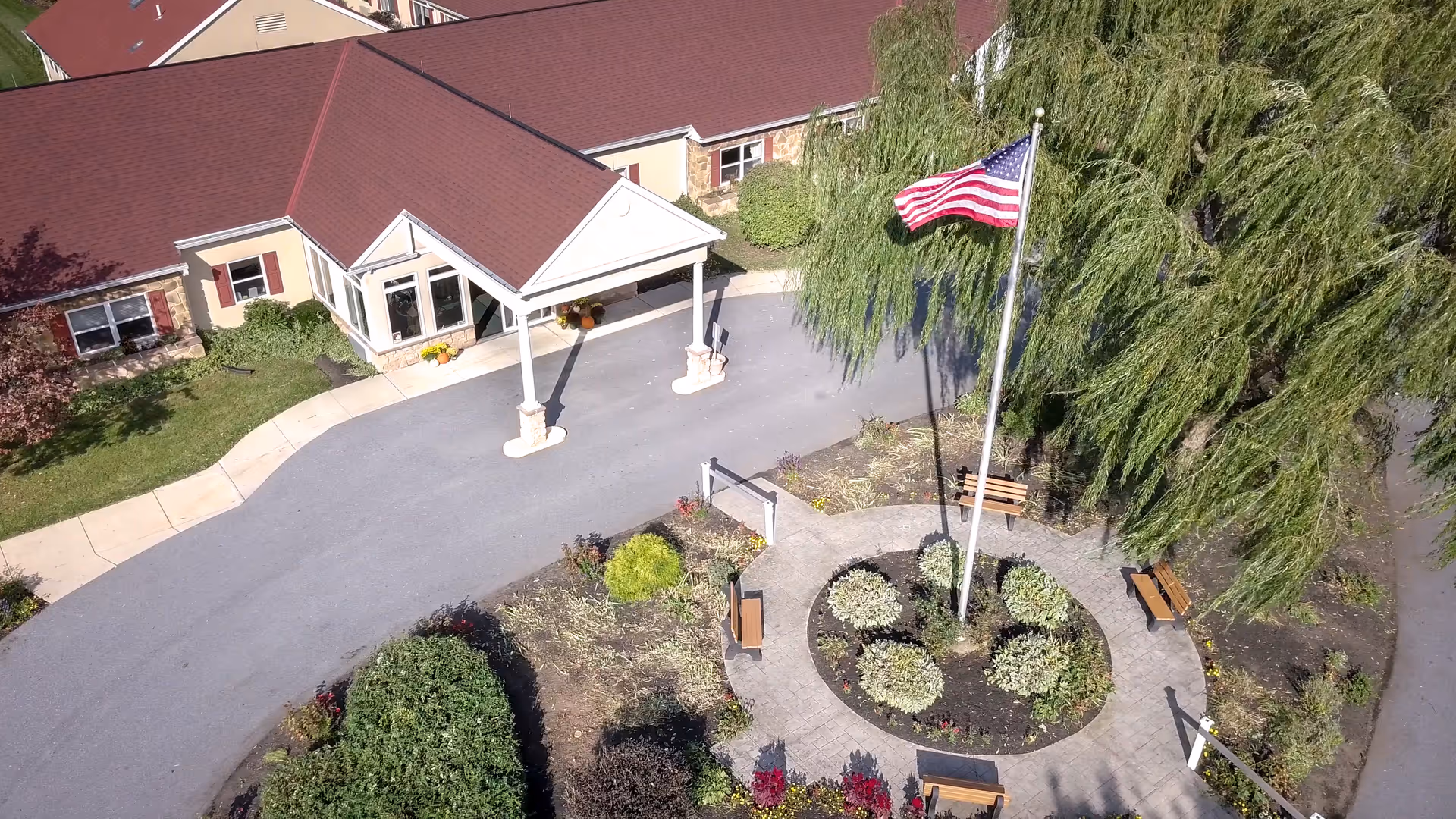 Aerial view of the entrance to Oak Leaf Manor South, showing a building with a red roof and beige walls. In front of the entrance is a circular garden with an American flag on a tall flagpole surrounded by benches and greenery. A paved driveway and sidewalks are also visible.