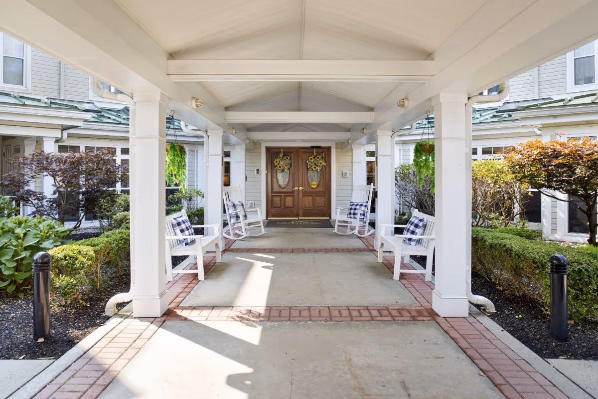 Covered front entrance with a walkway and white pillars leading to double wooden doors flanked by rocking chairs and greenery.