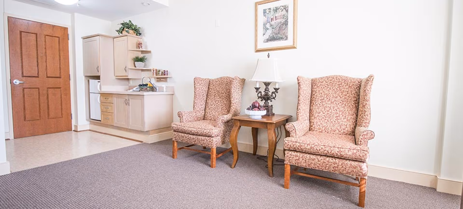 A cozy interior room featuring two patterned wingback chairs separated by a wooden side table with a decorative lamp and a bowl of fruit. In the background, there is a small kitchenette with light-colored cabinets, a sink, and a refrigerator. A wooden door is visible to the left, and a framed picture hangs on the wall above the chairs.