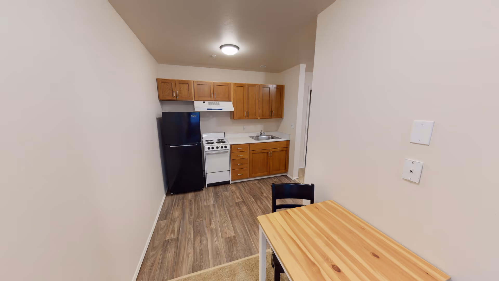 Small kitchen area with wooden cabinets, a black refrigerator, a white stove with an overhead vent, a sink, and a wooden table with a black chair in the foreground. The floor has wood-like vinyl planks and the walls are painted beige.