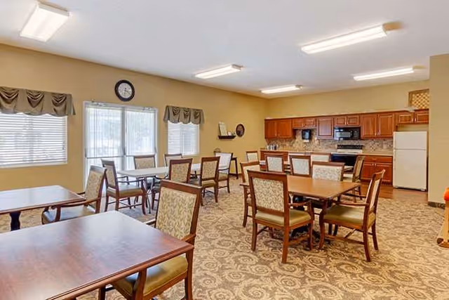 A dining room with multiple wooden tables and cushioned chairs arranged neatly. The room has beige walls, patterned carpet flooring, and large windows with valances allowing natural light. In the background, there is a kitchen area with wooden cabinets, a refrigerator, microwave, and stove.