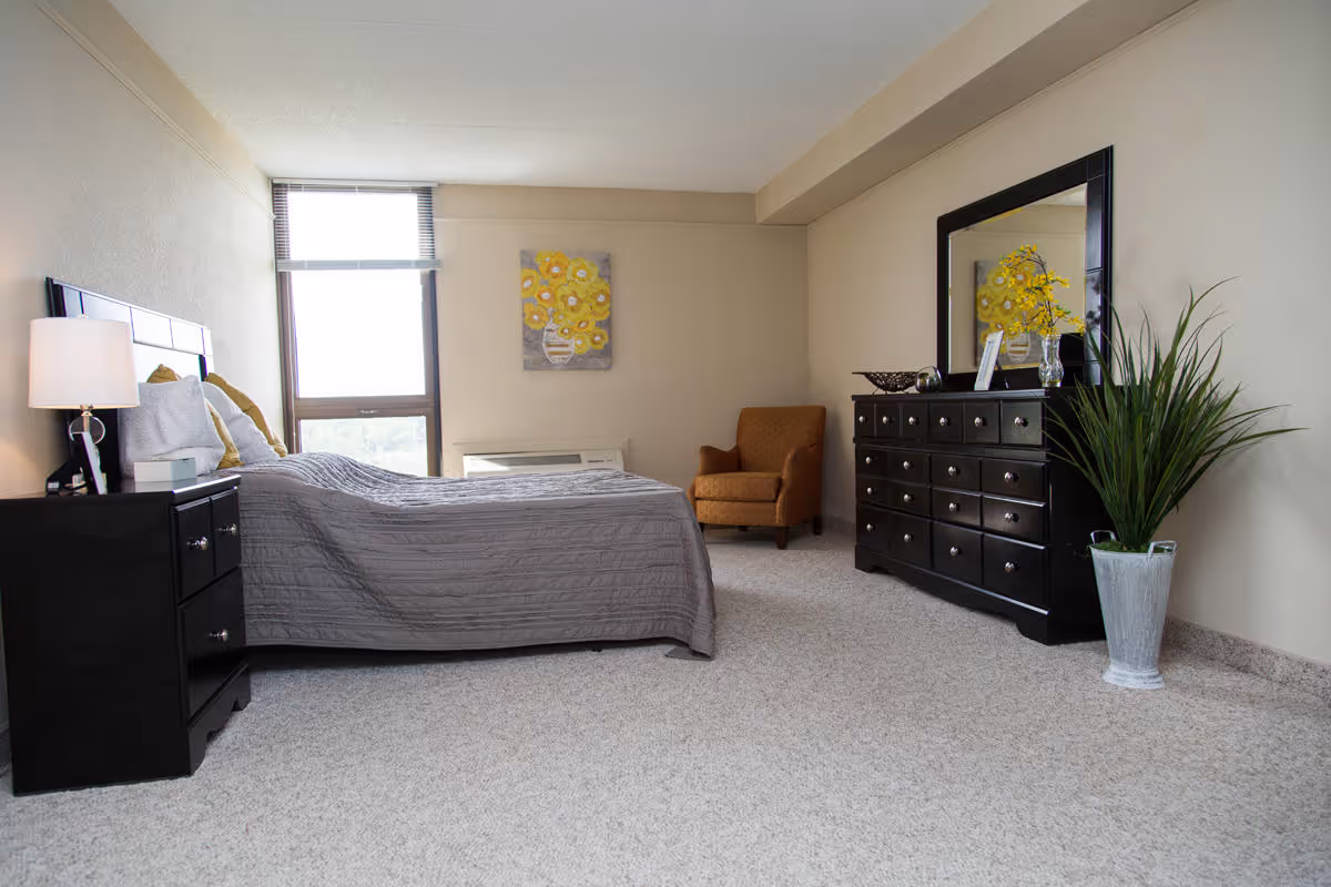 A bright and spacious bedroom with a large window letting in natural light. The room features a bed with gray bedding, a black nightstand with a lamp, a black dresser with a large mirror, a potted plant, a yellow floral painting on the wall, and a comfortable brown armchair in the corner.