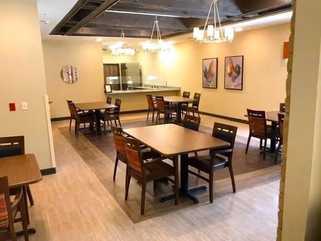 A dining room with several wooden tables and chairs arranged neatly on a wood and tile floor. The room has beige walls with two framed artworks and modern chandeliers hanging from the ceiling. There is a counter area with a sink in the background.