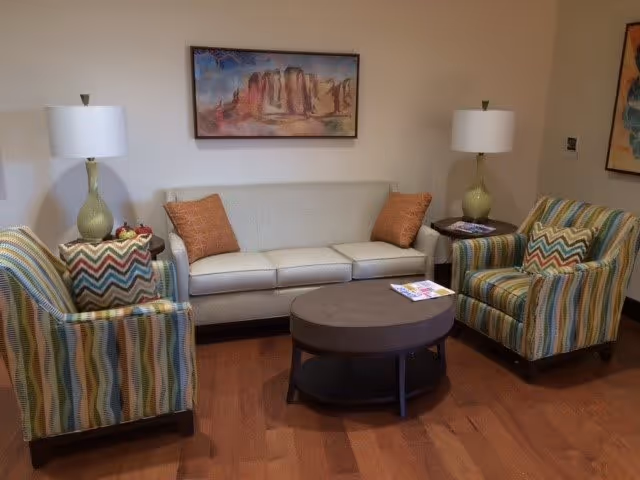 A cozy living room area with a white sofa adorned with two orange pillows, flanked by two colorful striped armchairs. A round dark coffee table sits in front of the sofa with a magazine on it. Two matching table lamps with green bases and white shades are placed on side tables next to each armchair. A framed landscape painting hangs on the beige wall above the sofa.