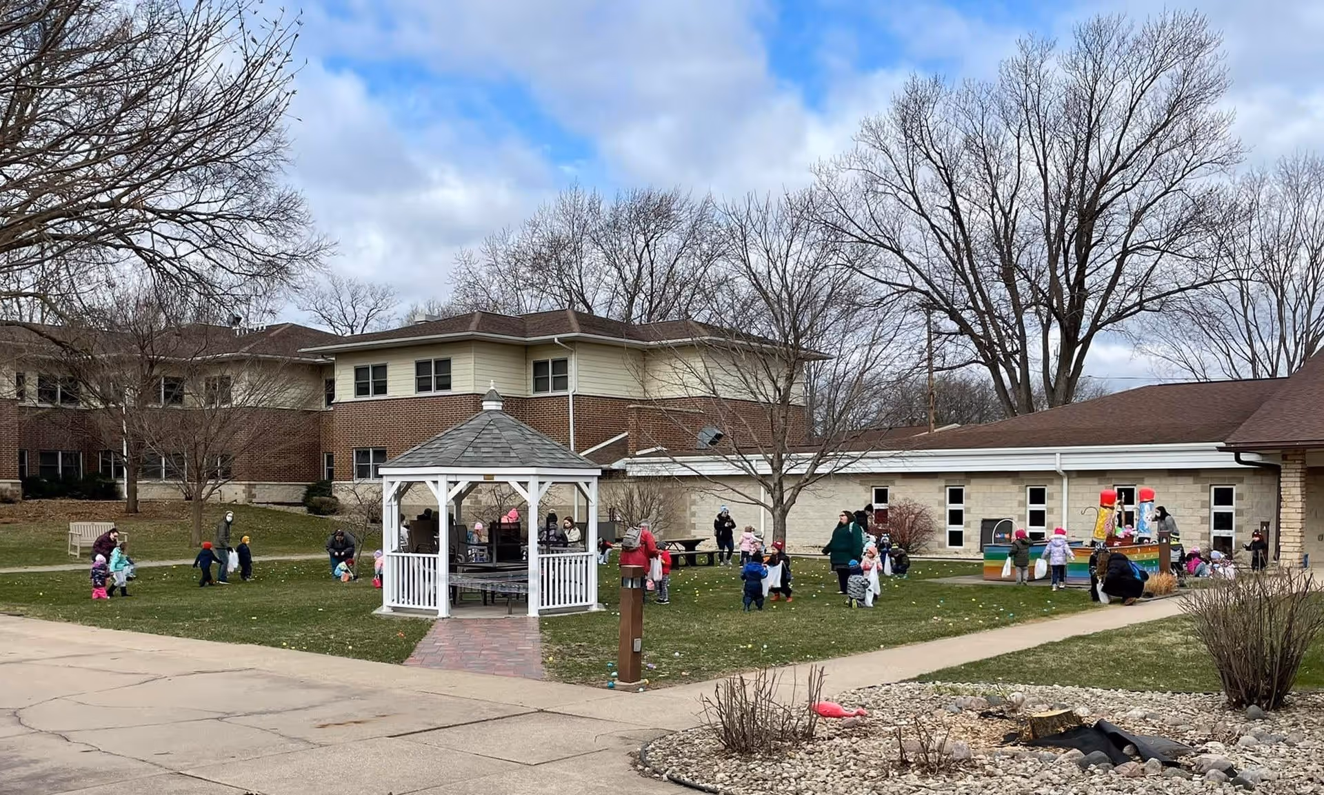 Outdoor grassy area at Benedictine Living Community La Crosse with a white gazebo in the center. Several children and adults are scattered around the lawn, some holding bags, likely participating in an Easter egg hunt. The background shows a two-story brick and beige building with leafless trees under a partly cloudy sky.