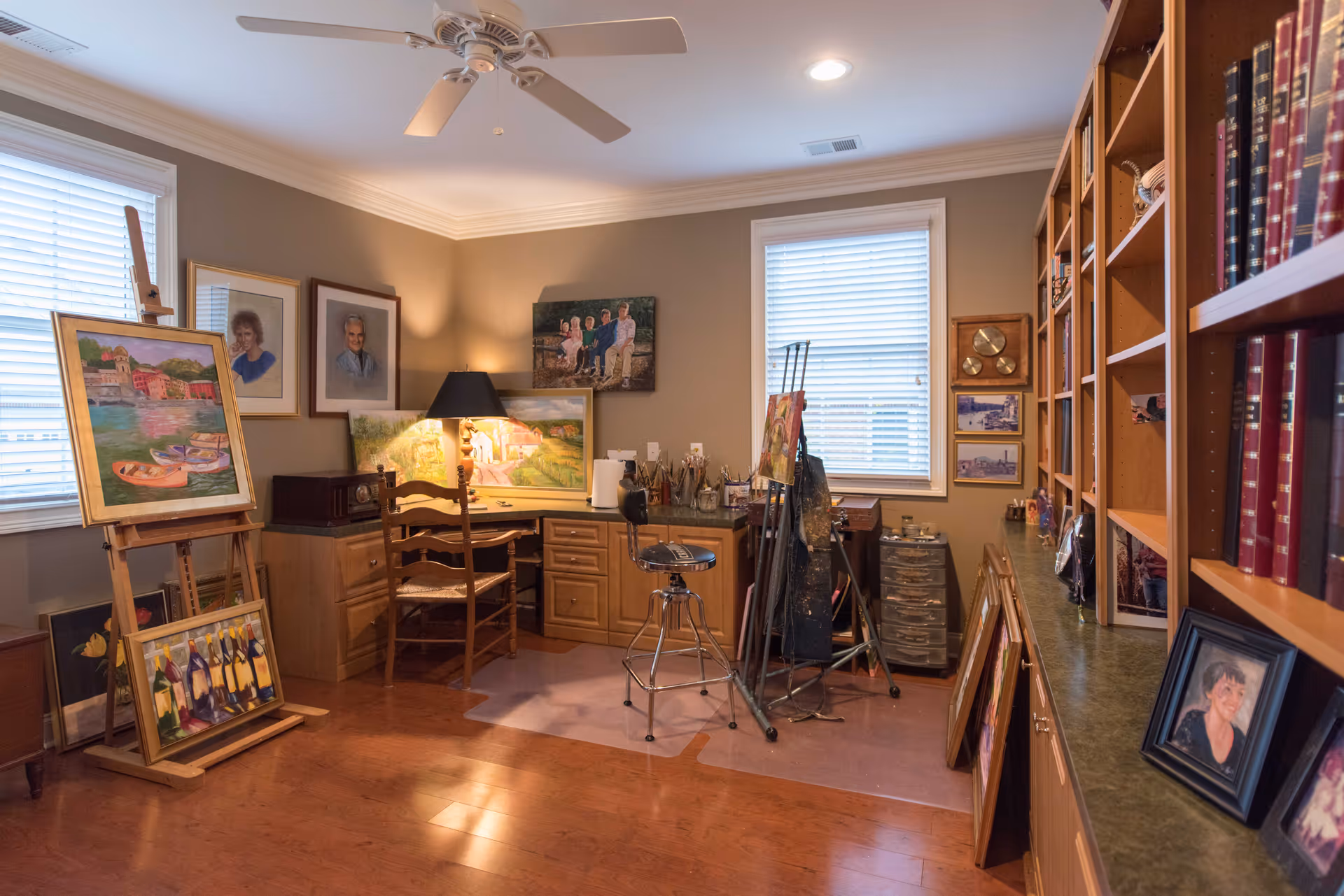 A cozy art studio room with wooden floors and beige walls. The room features a wooden desk with a chair, a lamp, and various painting supplies. Several paintings and portraits are displayed on easels and walls. There are two windows with blinds, a ceiling fan, and built-in wooden bookshelves filled with books and framed photos.
