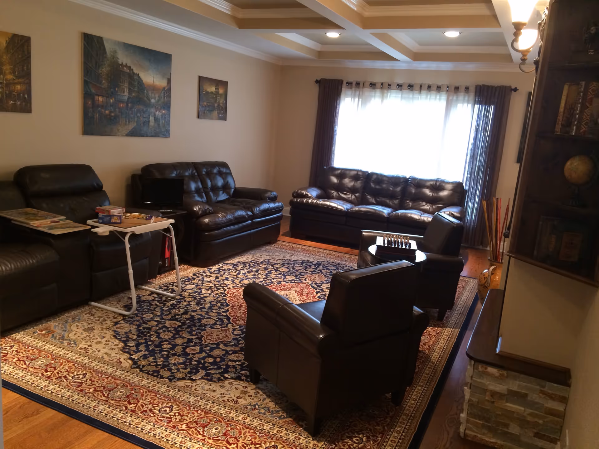 A cozy living room with multiple dark leather sofas and armchairs arranged around a large patterned area rug. The room features a window with sheer curtains and darker drapes, paintings on the wall, a small table with board games, and a bookshelf with books and a globe.