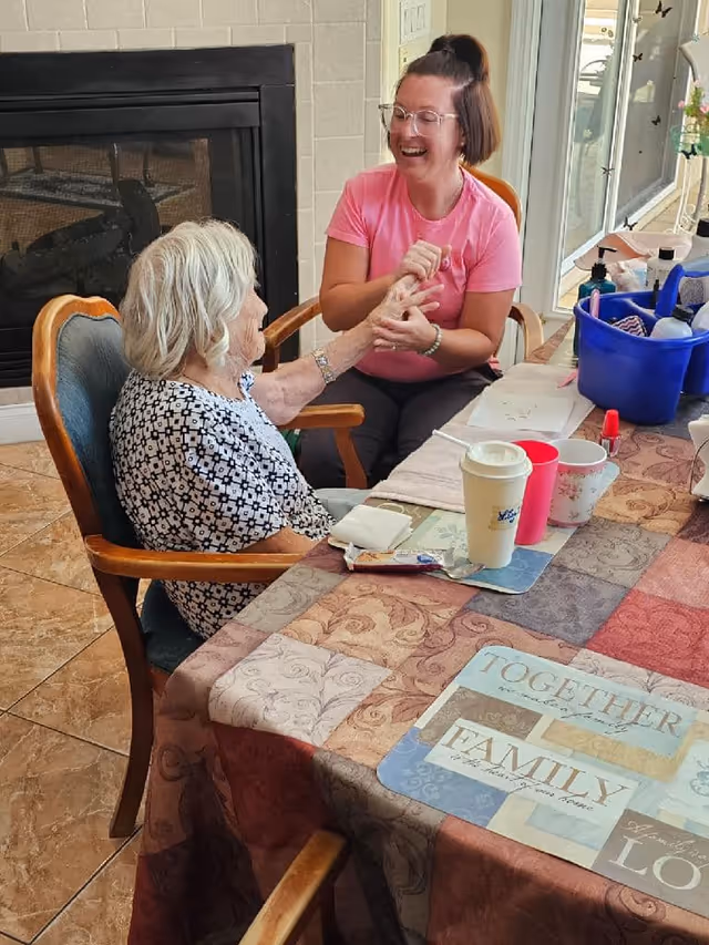 An elderly woman with white hair sitting at a table covered with a patterned tablecloth, holding hands and smiling with a younger woman wearing glasses and a pink shirt. The setting appears to be a cozy indoor room with a fireplace in the background and a large window or glass door letting in natural light. Various cups and a blue container with items are on the table.