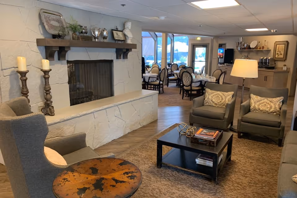 A cozy senior living facility common area featuring a stone fireplace with decorative items on the mantel, two armchairs with patterned pillows, a coffee table with books and decorative objects, and a round side table. In the background, there is a dining area with multiple tables and chairs, large windows letting in natural light, and a beverage station with coffee dispensers and cups.