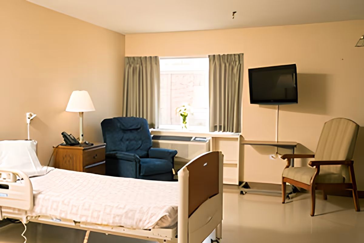 A senior care patient room with a hospital bed, a blue recliner, side table with lamp and phone, a wall-mounted TV and a chair by a window.