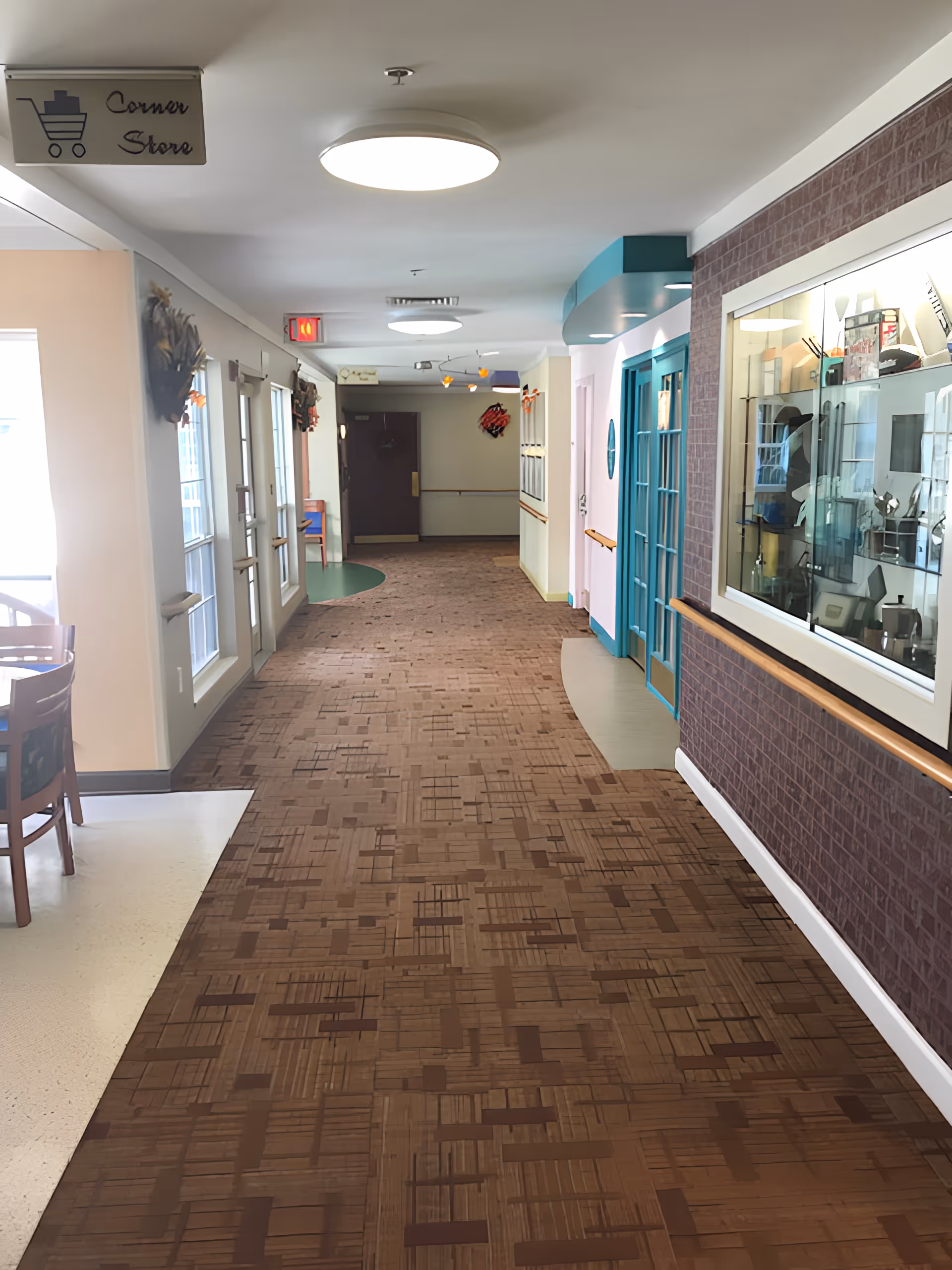 A hallway in a senior living facility with patterned brown carpet and white walls. On the left side, there are windows and a dining area with wooden chairs and tables. A sign hanging from the ceiling reads 'Corner Store' with a shopping cart icon. On the right side, there is a display case with various items and a blue door. The hallway is well-lit with ceiling lights and has handrails along the walls.