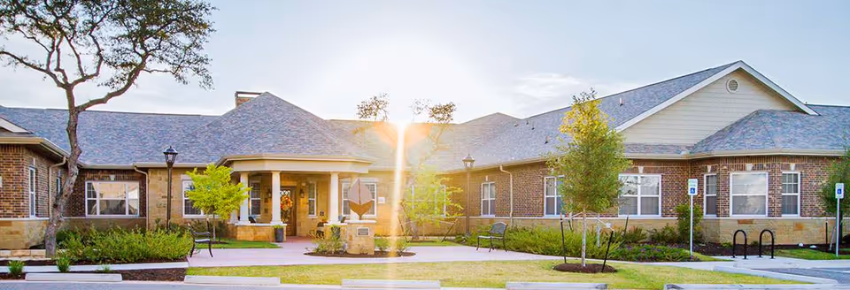 Exterior view of Silverado Barton Springs Memory Care Community building with brick walls, multiple windows, a covered entrance with columns, landscaped greenery, benches, and a clear sky with sunlight shining.