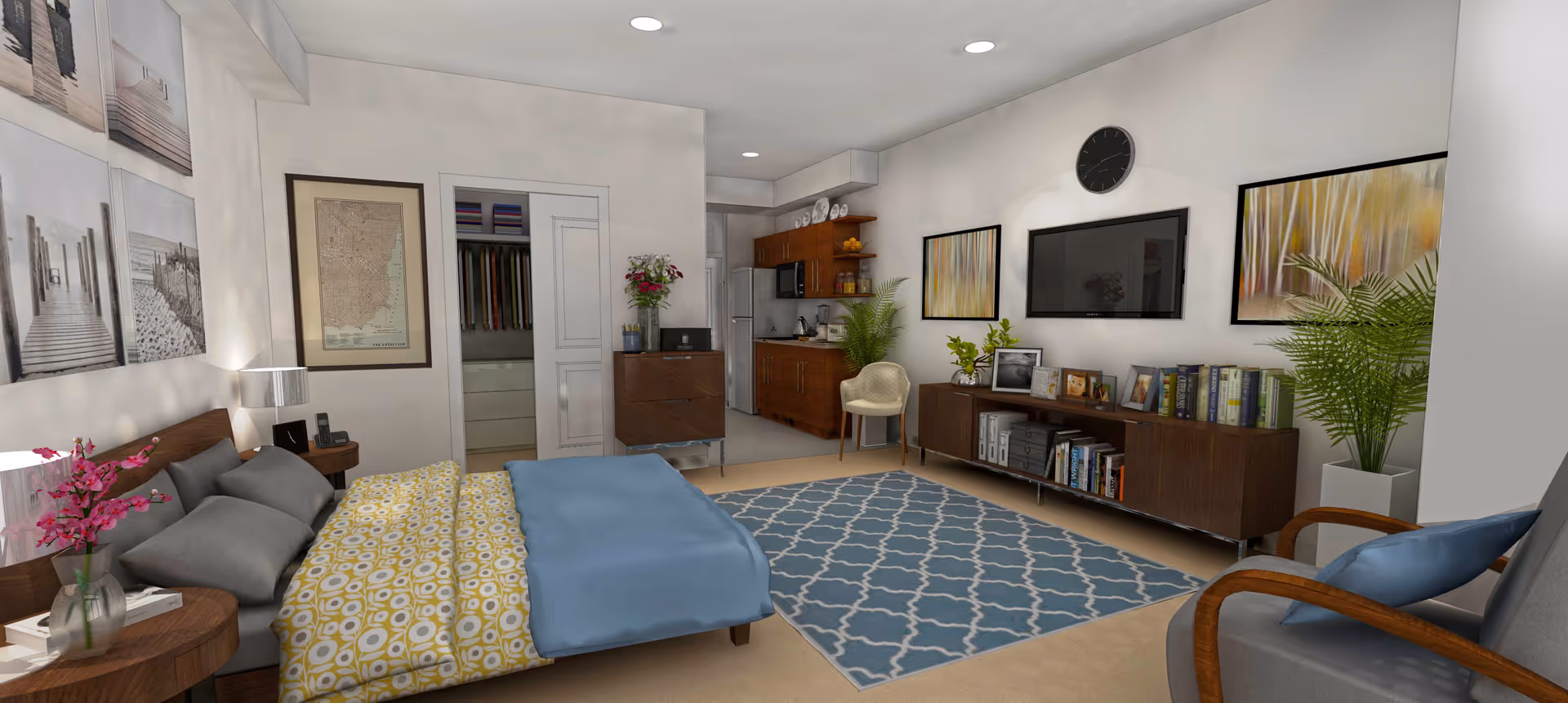 A modern and cozy bedroom with a bed featuring gray pillows and a yellow patterned blanket with a blue throw. There are two wooden nightstands with lamps and a vase of pink flowers. The room has a blue patterned rug, a wooden chair with a blue cushion, and a wooden cabinet with books and framed photos. A flat-screen TV and two abstract paintings hang on the wall above the cabinet. A small kitchenette with wooden cabinets, a microwave, and a refrigerator is visible in the background. There is also a closet with sliding doors and a vase with flowers on a small dresser.