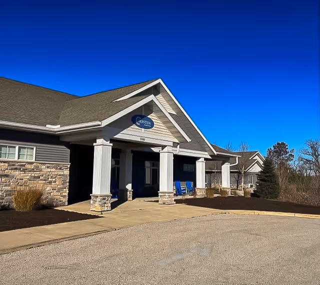 Front exterior view of Frontida Assisted Living of Elkhorn building with a covered entrance supported by white columns, stone and siding facade, blue chairs on the porch, and clear blue sky above.
