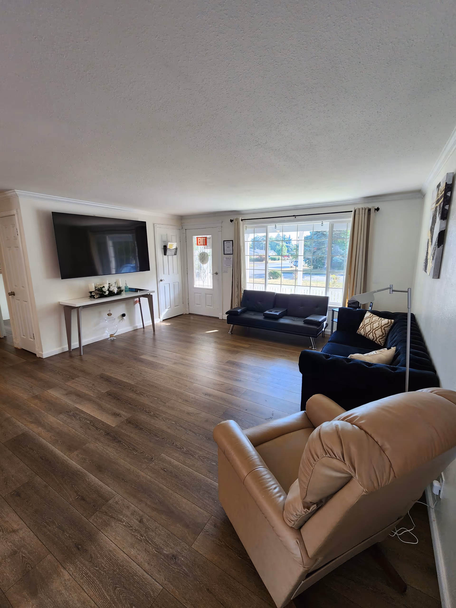 A bright living room with wood flooring, featuring a beige recliner chair in the foreground, two dark sofas near a large window with beige curtains, a wall-mounted flat-screen TV above a small table with decorative items, and a door with an exit sign.