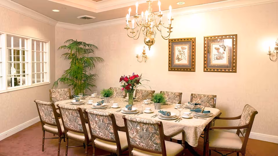 A formal dining room with a long rectangular table covered with a beige tablecloth, set with plates, cups, napkins, and silverware. The table is surrounded by ten upholstered chairs with floral patterns. A chandelier hangs above the table, and two framed butterfly-themed artworks are on the wall. There is a large green potted plant in the corner and a window with multiple panes on the left wall.