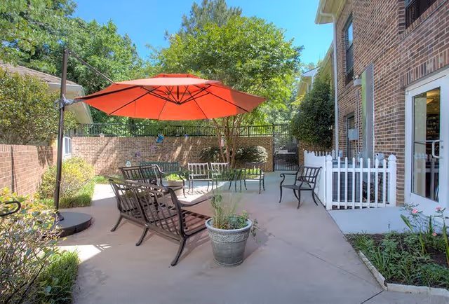 Outdoor patio area with metal benches and chairs arranged around a large red umbrella. The patio is surrounded by brick walls and greenery, with a white picket fence section near a building entrance door.