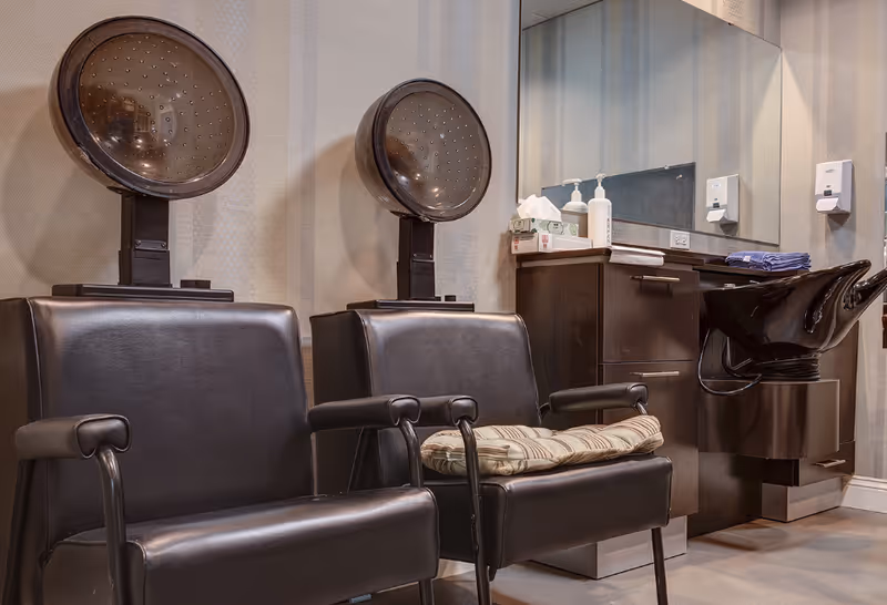 Salon area with two hooded hair dryers over black leather chairs, a shampoo sink, mirror, and cabinetry.