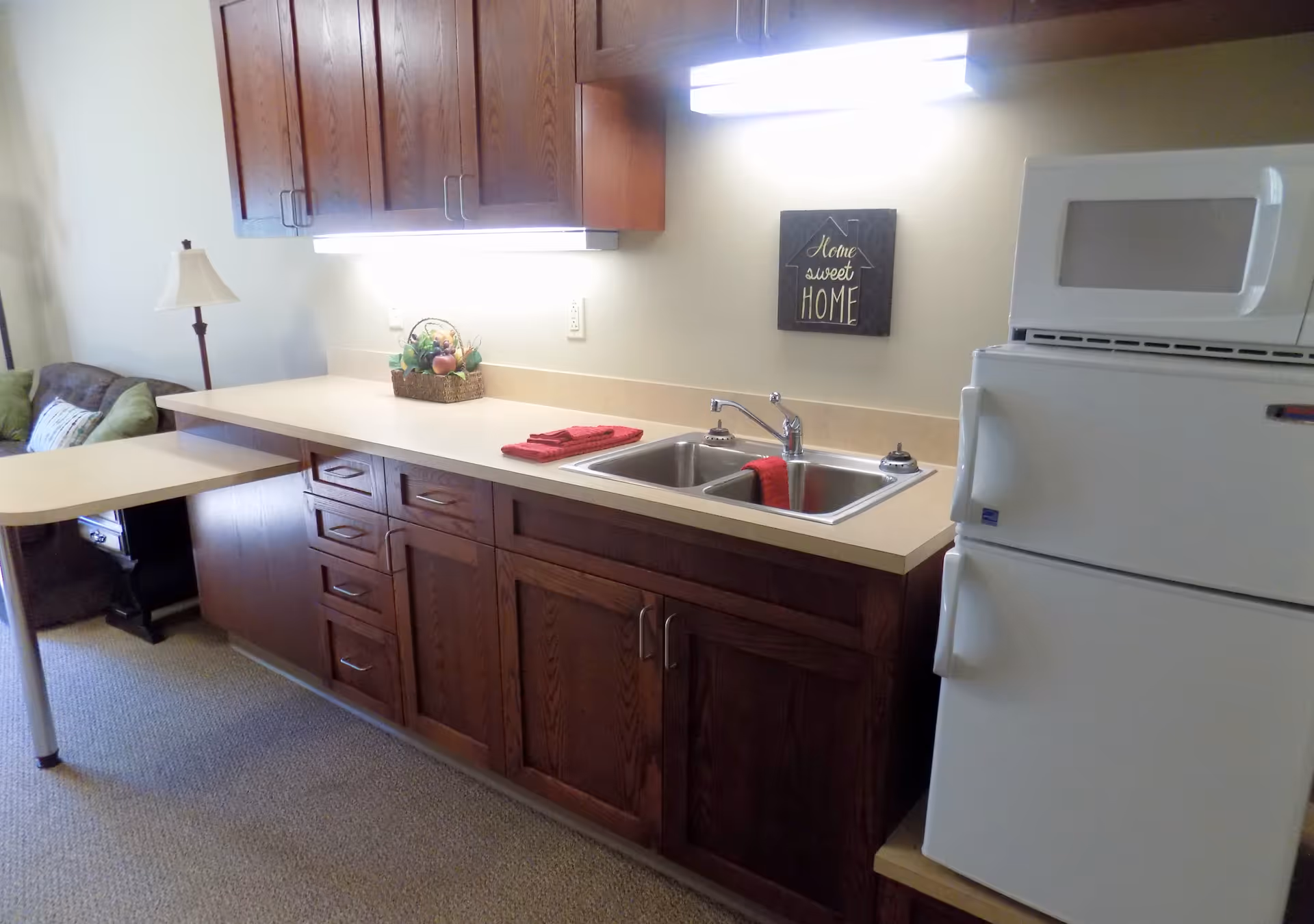 A kitchenette area with wooden cabinets, a double sink with a red towel, a white microwave on top of a white refrigerator, and a countertop with a basket of fruit and red towels. In the background, there is a small table attached to the counter and a green couch with a lamp beside it. A wall decoration reads 'Home sweet HOME'.