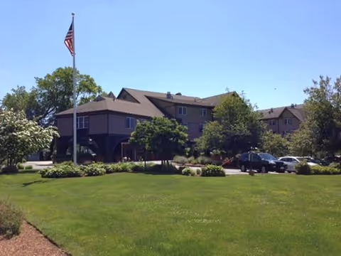 Front exterior of a multi-story senior living building with a flagpole, landscaped lawn, and parked cars.