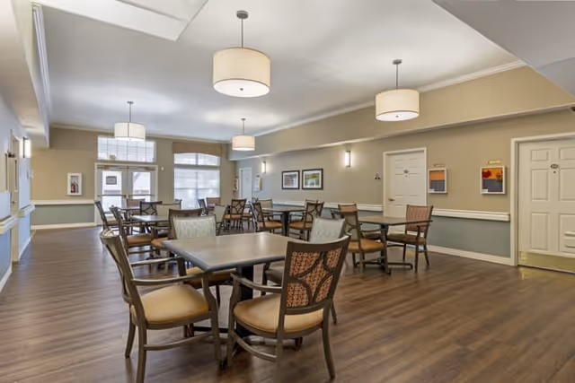 A spacious dining room with multiple tables and chairs arranged neatly on a wooden floor. The room has beige walls with white trim, several framed pictures on the walls, and large windows letting in natural light. Ceiling lights with round fabric shades hang evenly spaced across the ceiling.