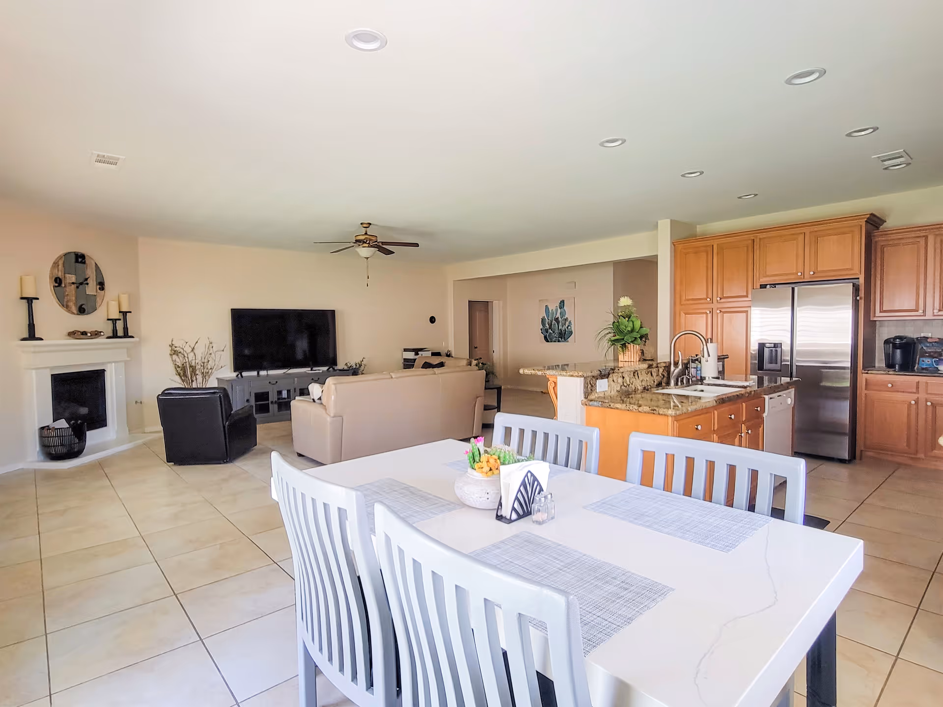 Open-plan interior with a dining table in the foreground, a living room with sofa and TV, and a kitchen with an island and stainless refrigerator.