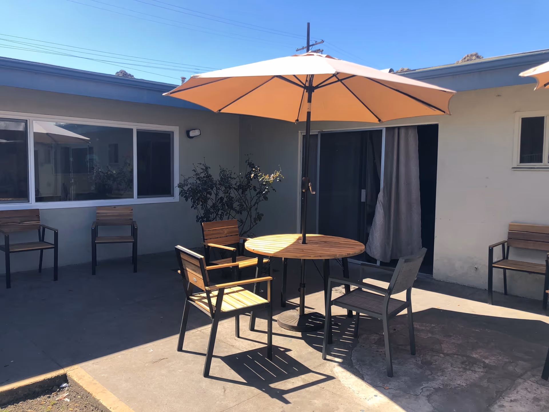 Outdoor patio area with a round wooden table and four chairs under a large beige umbrella. The patio is adjacent to a building with windows and a sliding glass door with curtains. Additional chairs are placed against the building walls.
