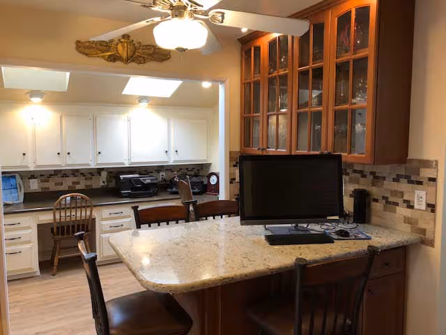 Interior view of a kitchen area featuring a granite countertop island with three chairs, a computer monitor on the island, wooden cabinets with glass doors, white cabinets along the back wall, and a ceiling fan with a light fixture.