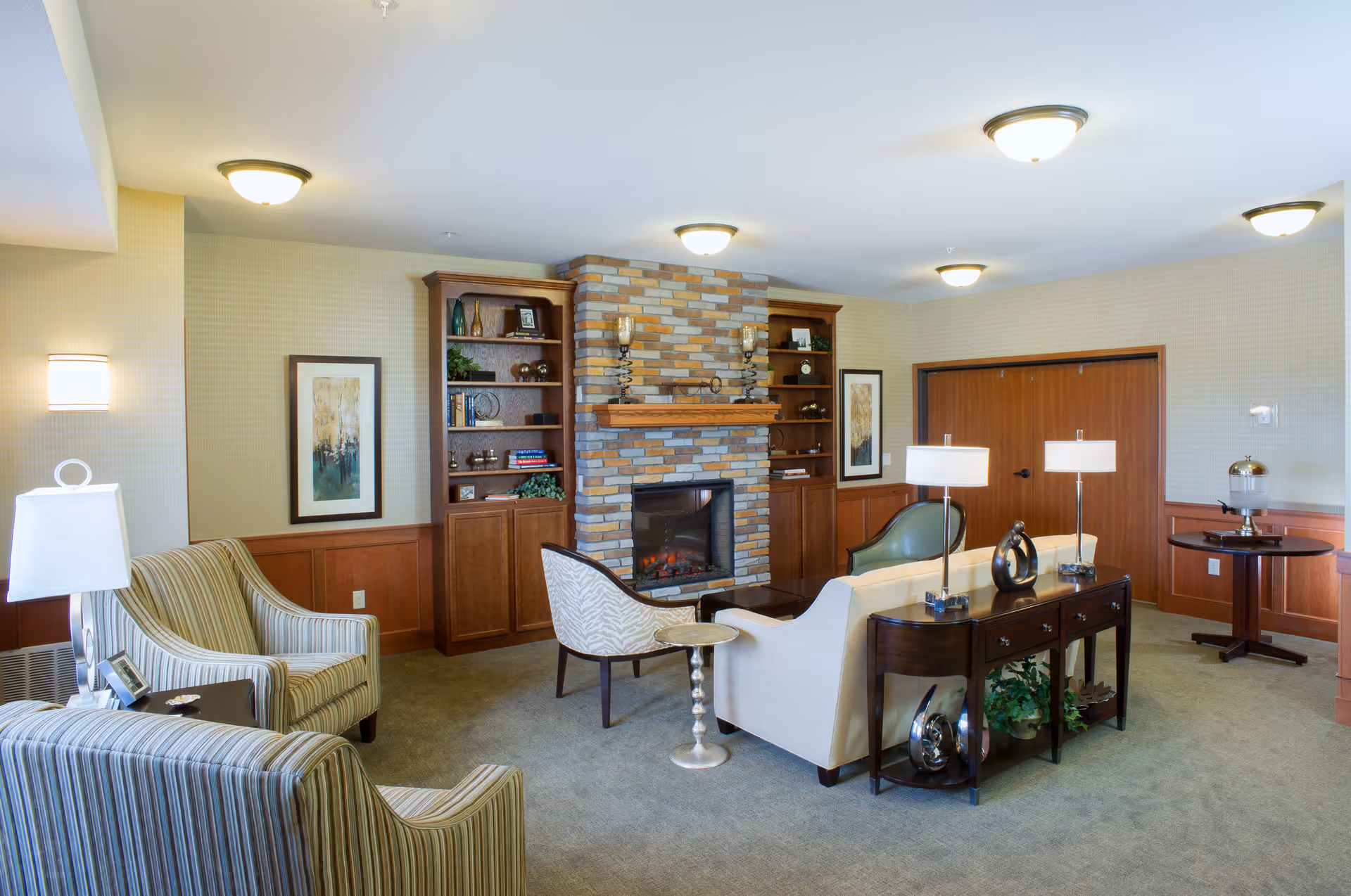 A cozy senior living facility common area featuring a stone fireplace flanked by wooden built-in shelves with decorative items and books. The room has comfortable seating including striped armchairs, a white sofa, and a patterned chair. There are two framed artworks on the walls, table lamps on a dark wooden console table behind the sofa, and a small round table with a beverage dispenser in the corner. The walls are light-colored with wood paneling on the lower half, and the ceiling has multiple flush-mounted lights.