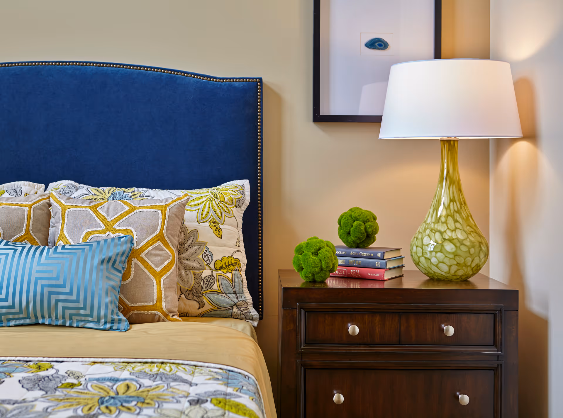 Close-up view of a bedroom corner featuring a bed with a blue upholstered headboard and multiple decorative pillows in blue, yellow, and beige patterns. Next to the bed is a dark wooden nightstand with two drawers, topped with a green patterned lamp, two green decorative moss balls, and a small stack of books. A framed artwork is partially visible on the wall above the nightstand.