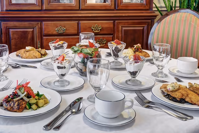 A dining table set with plates of food including grilled chicken with vegetables, fried fish with sides, and dessert parfaits topped with strawberries. The table is covered with a white tablecloth and set with glassware, cups, and silverware. In the background, there is a wooden cabinet and a striped upholstered chair.