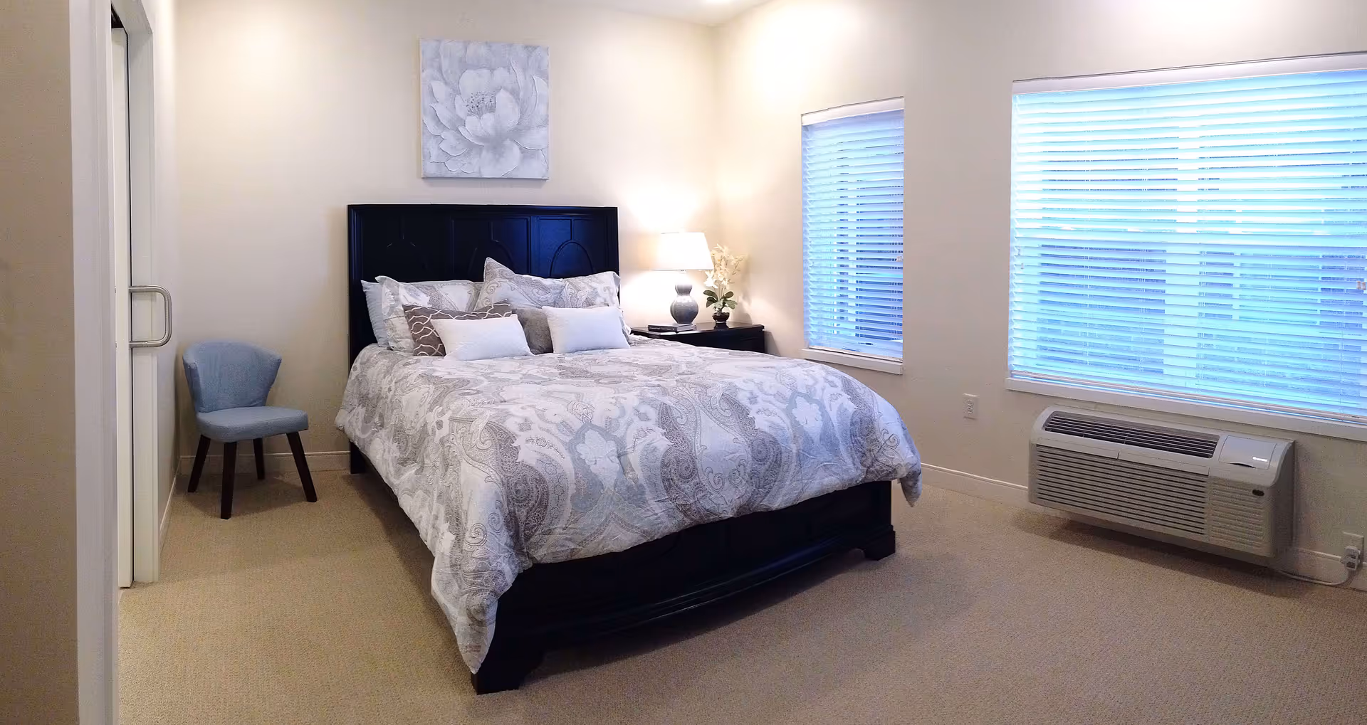 Bright bedroom with a patterned bed and dark headboard, nightstand and lamp, a blue chair, and two large windows with blinds.