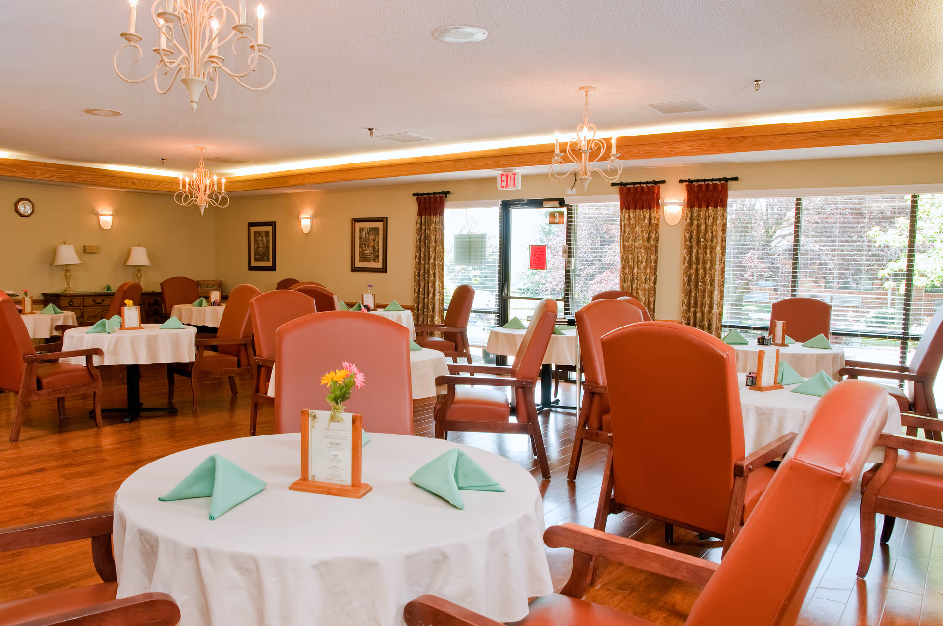 A bright dining room with round tables covered in white tablecloths, each set with green folded napkins and a small flower arrangement. The room has wooden floors, red cushioned chairs, chandeliers, wall lamps, framed artwork, and large windows with curtains letting in natural light.