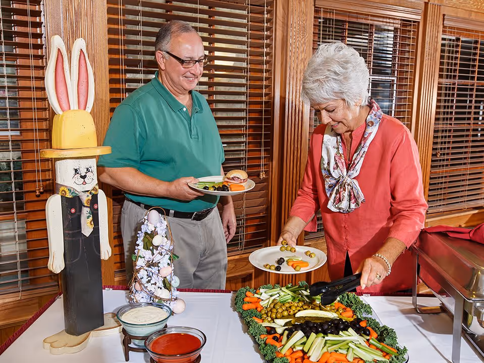 An elderly man and woman serving themselves food from a buffet table inside a room with wooden blinds. The man is holding a plate with a sandwich and some olives, while the woman is using tongs to pick vegetables from a tray. There is a decorative wooden bunny figure and an Easter-themed decoration on the table.