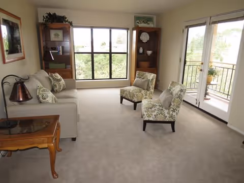 A bright living room with beige carpet, a light-colored sofa with patterned cushions, two patterned armchairs, a wooden side table with a lamp, two wooden bookshelves with decorative items, a large window overlooking greenery, and glass doors leading to a balcony.
