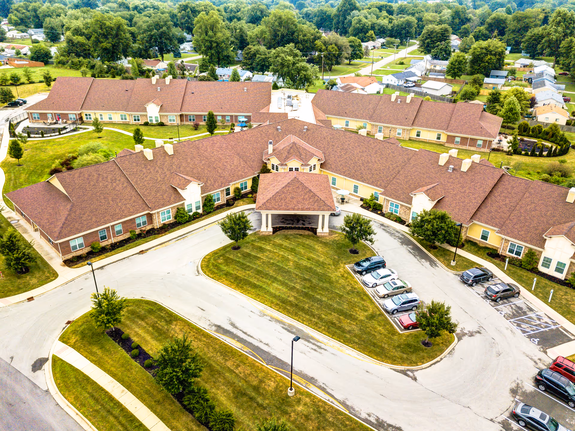 Aerial view of Symphony at Valley Farms senior living facility showing a large, single-story building with a brown roof and beige walls. The building is surrounded by green lawns, trees, and a parking lot with several cars. Residential houses and trees are visible in the background.