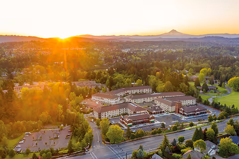 Aerial view of Kellogg Assisted Living At Mary's Woods facility at sunset, surrounded by trees and greenery with a mountain visible in the background and parking lots adjacent to the buildings.