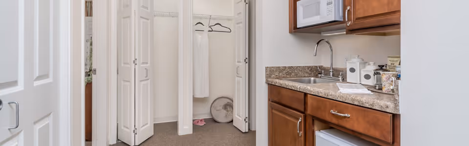Interior view of a senior living facility unit showing a small kitchenette with a sink, countertop, microwave, and wooden cabinets. To the left, there is an open closet with a white dress hanging and a pair of pink slippers on the floor. The walls are painted white and the space appears clean and well-lit.