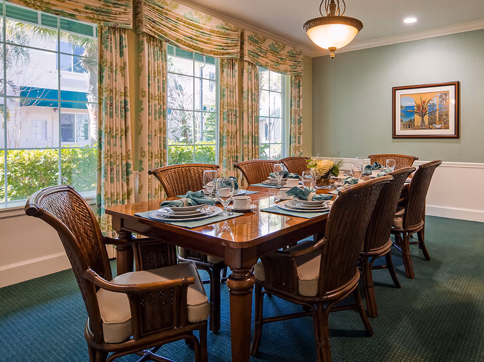 Formal dining room with a set wooden table and wicker chairs beside large windows with floral curtains.