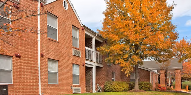 Exterior view of a brick senior living facility building with multiple windows and a balcony. There are trees with orange autumn leaves and well-maintained bushes in front of the building under a partly cloudy sky.