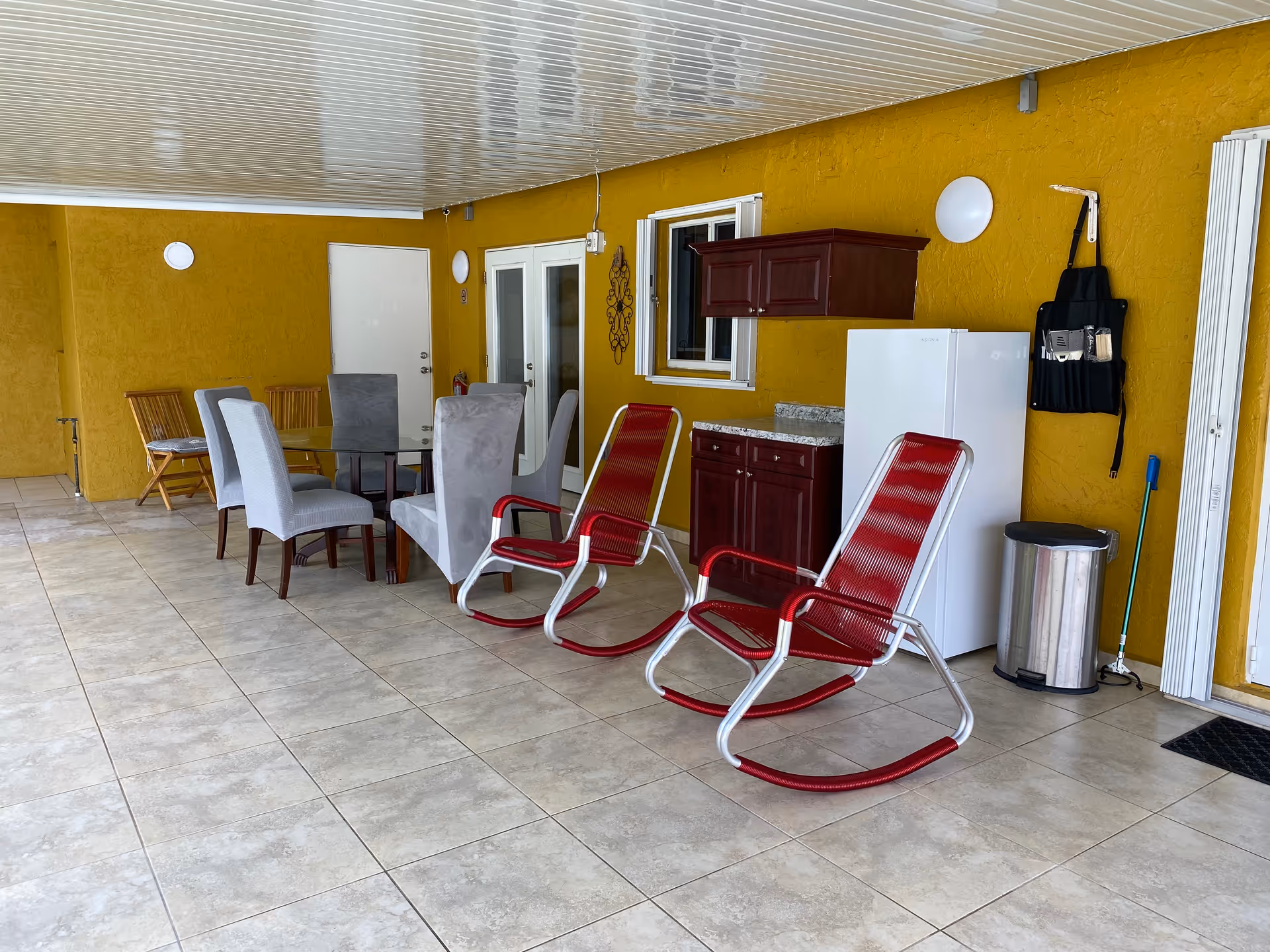 Covered seating area with a dining table and upholstered chairs, two red rocking chairs, and a small kitchenette with cabinets and a refrigerator against a yellow wall.